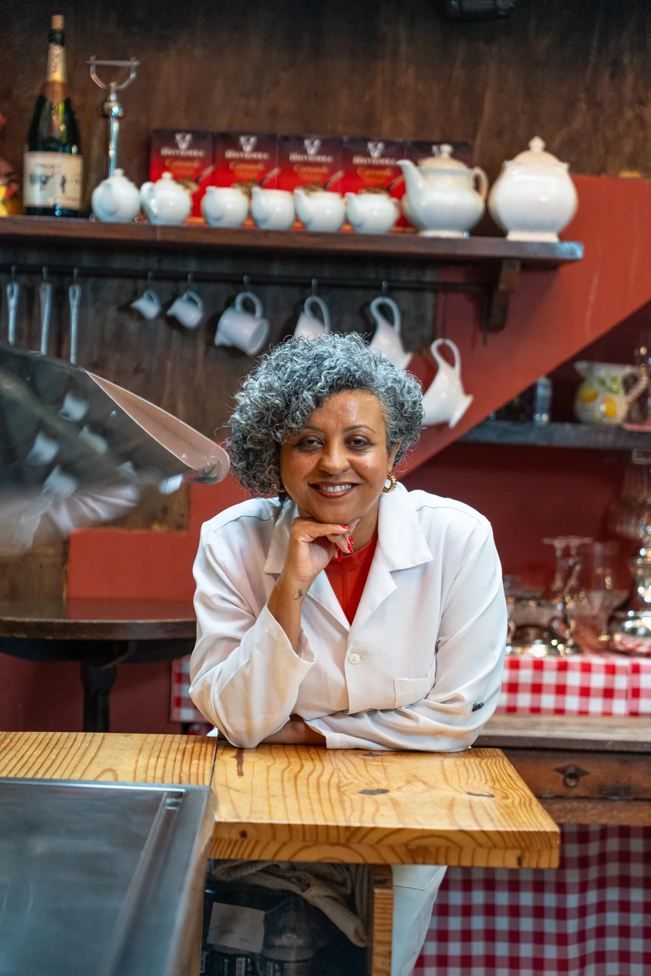 Tatiana Queiroz de jaleco branco em uma cozinha, apoiada em uma mesa de madeira.